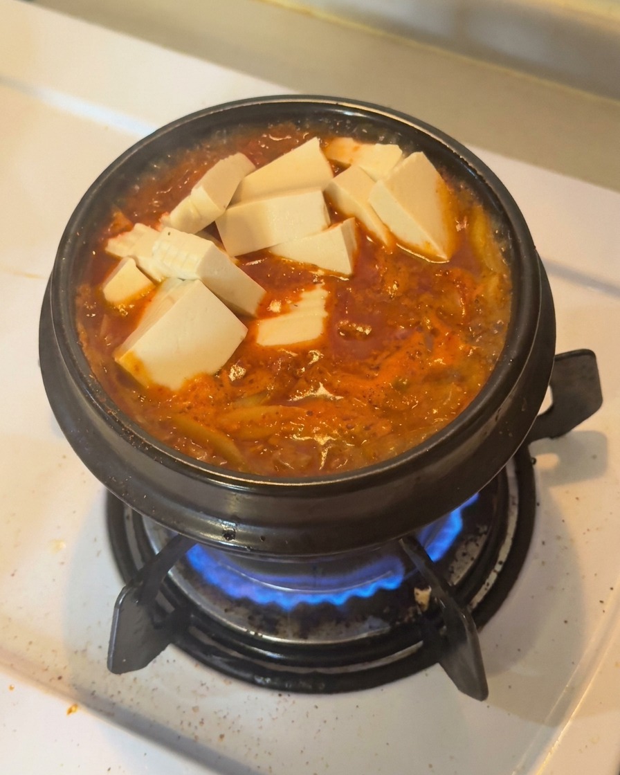 Fresh white tofu cubes and sliced green chilies arranged on top of a bubbling red kimchi stew in a black earthenware pot.