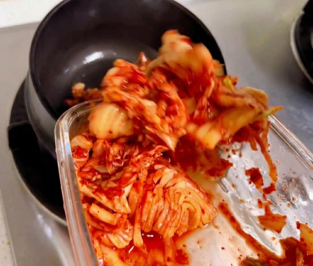 Chopped kimchi being stirred and sautéed in a traditional black stone pot over a gas flame.