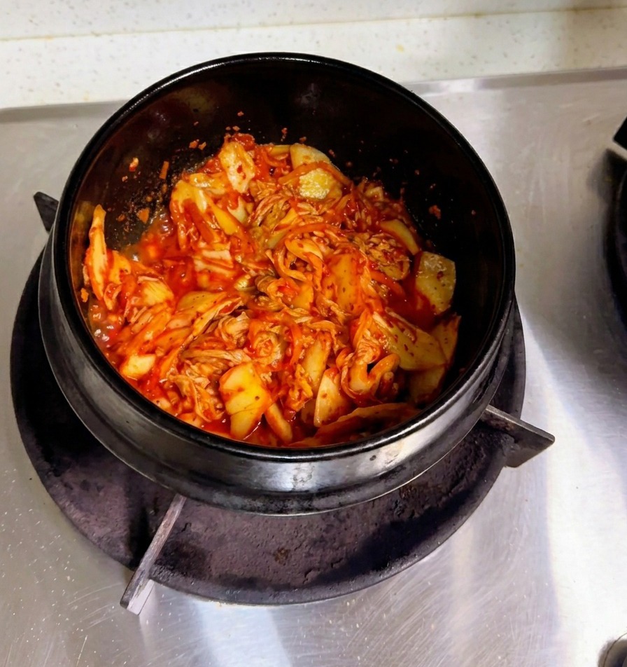 A hand pouring cloudy rice water into a stone pot filled with sautéed kimchi.