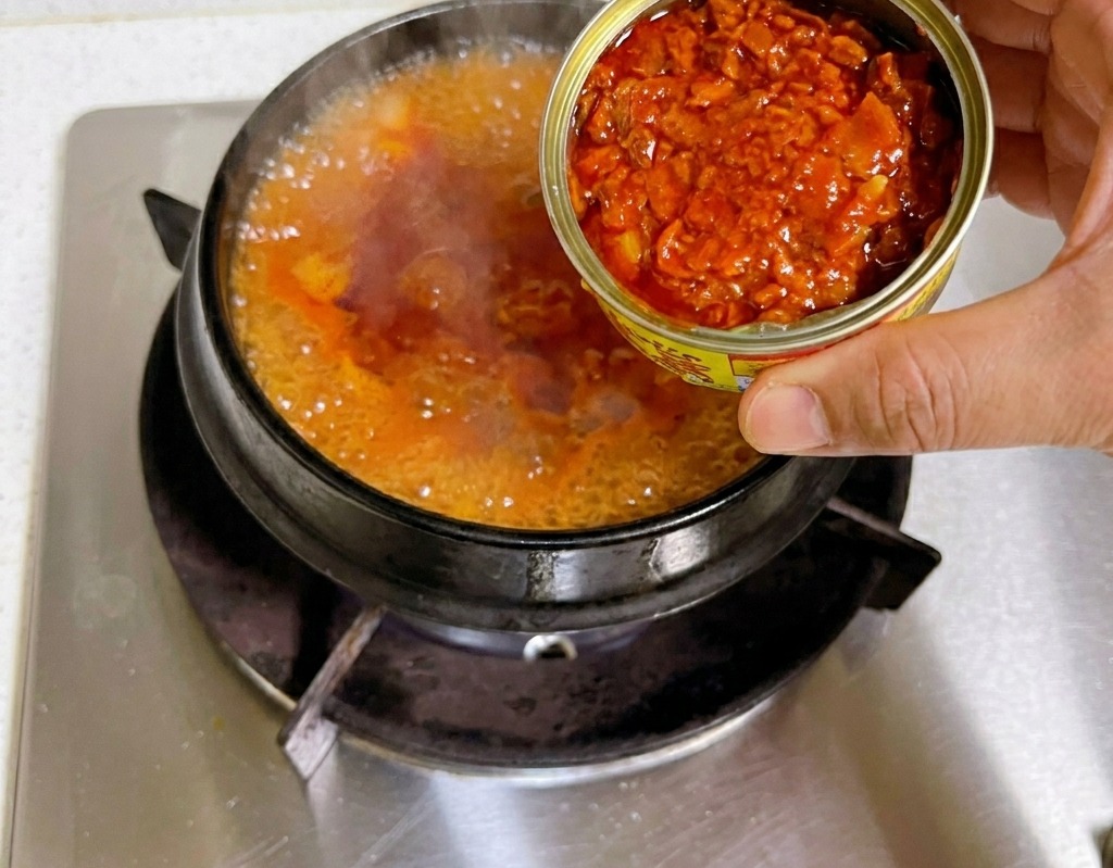 A hand holding an open tin can and pouring chunks of tuna into a boiling red kimchi stew base in a stone pot.