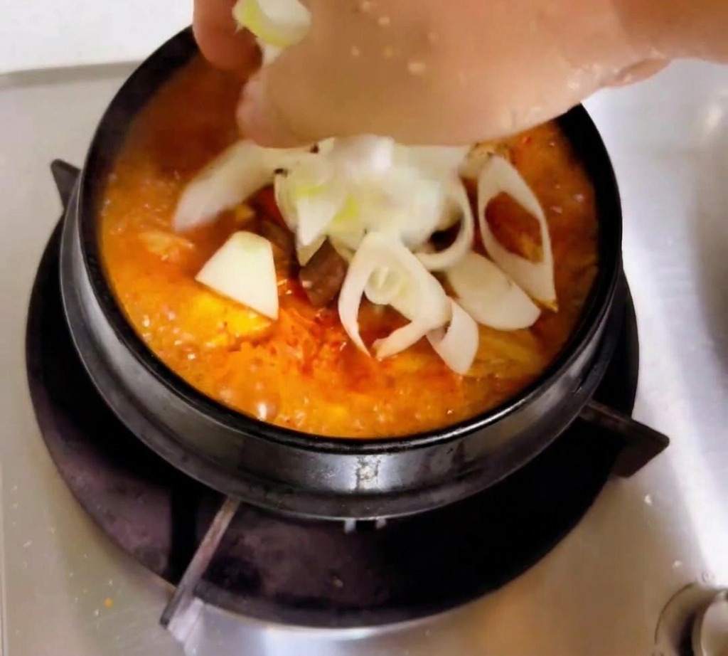 A hand adding sliced white onions and scallions to a simmering orange-red kimchi stew.