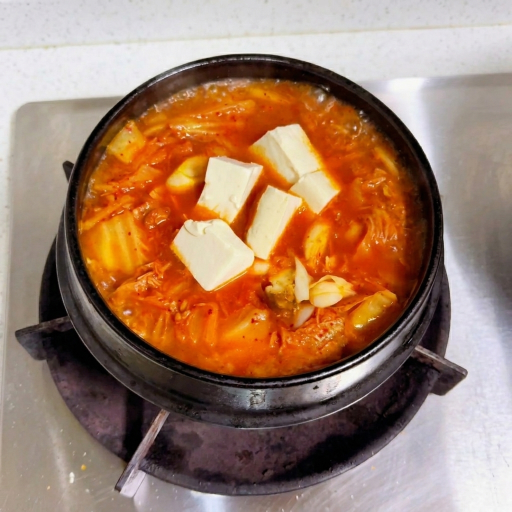 Cubes of fresh white tofu resting on top of a bubbling red kimchi broth in a black pot.
