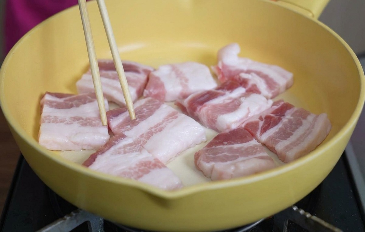 Raw thick slices of pork belly laid out in a yellow ceramic frying pan, being adjusted with chopsticks.