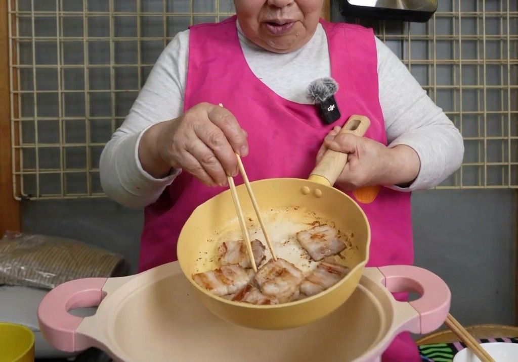 Slices of browned pork belly being moved with chopsticks from a yellow skillet into a larger pink pot.