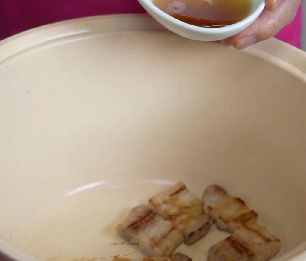 A small white bowl pouring golden sesame oil over fried pork belly pieces in a large cream-colored pot.