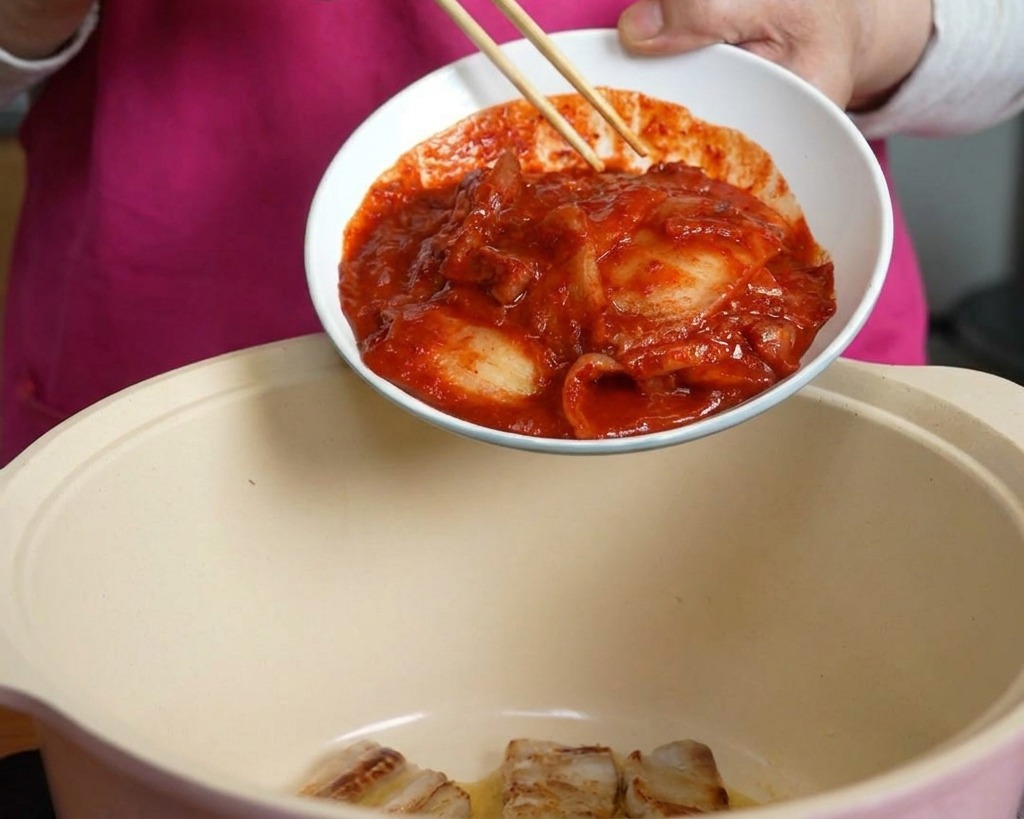 A hand using chopsticks to slide a bowl of spicy red kimchi into a cooking pot containing pork belly.