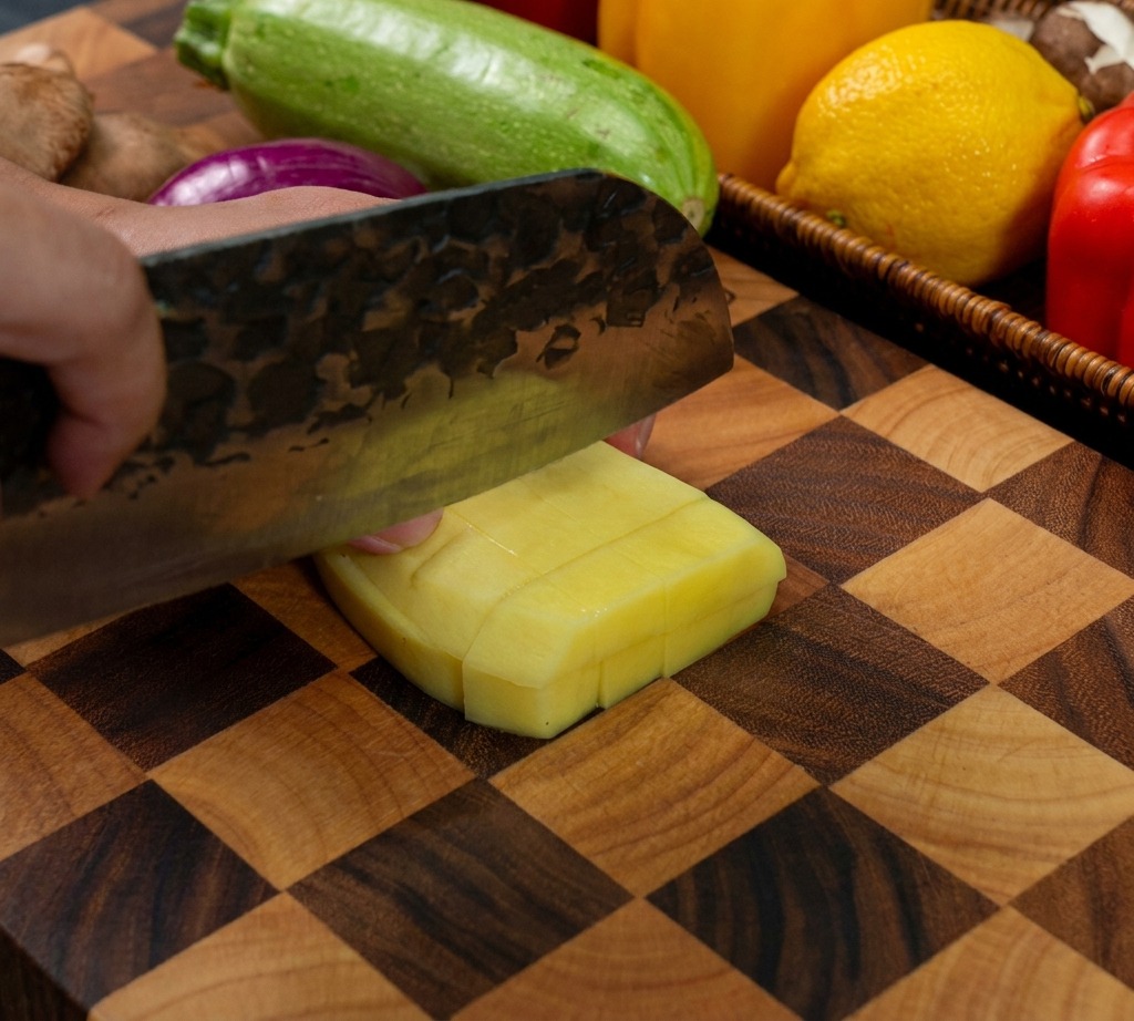 A heavy-duty knife slicing through a peeled yellow potato to create small, even cubes on a wooden cutting board.