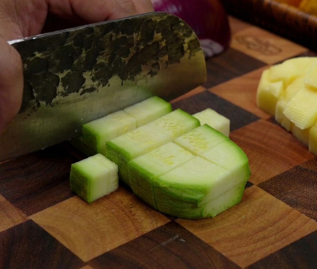 A cleaver cutting through a green zucchini, dividing it into small rectangular chunks on a checkered wood board.