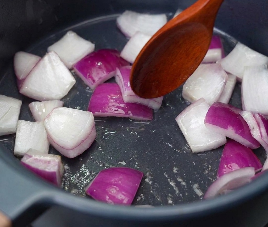 Chopped red onion pieces being stirred with a wooden spoon in a dark pot over medium heat.