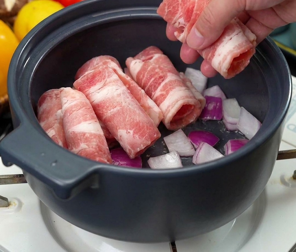 A hand placing several rolls of thinly sliced raw beef into a pot over a bed of sautéing onions.