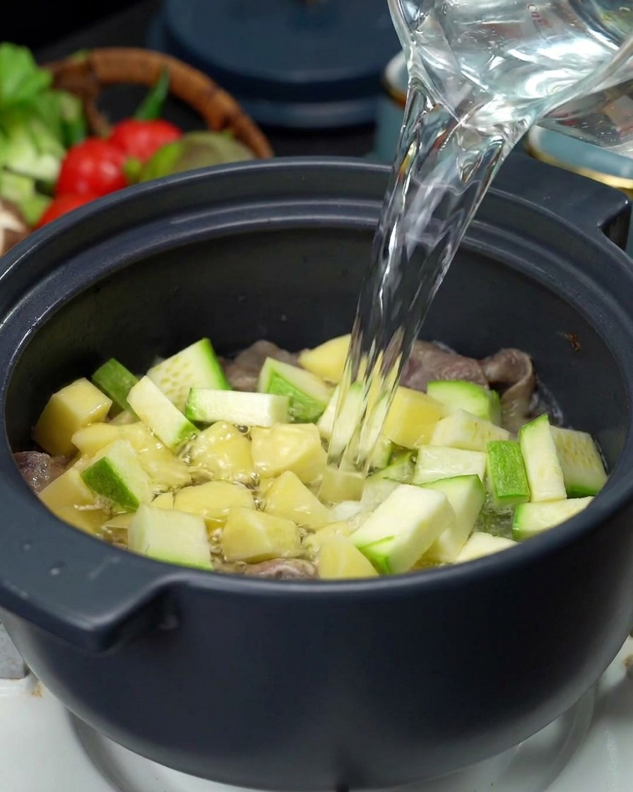 A stream of clear water being poured from a glass pitcher into a pot filled with meat, potatoes, and zucchini.