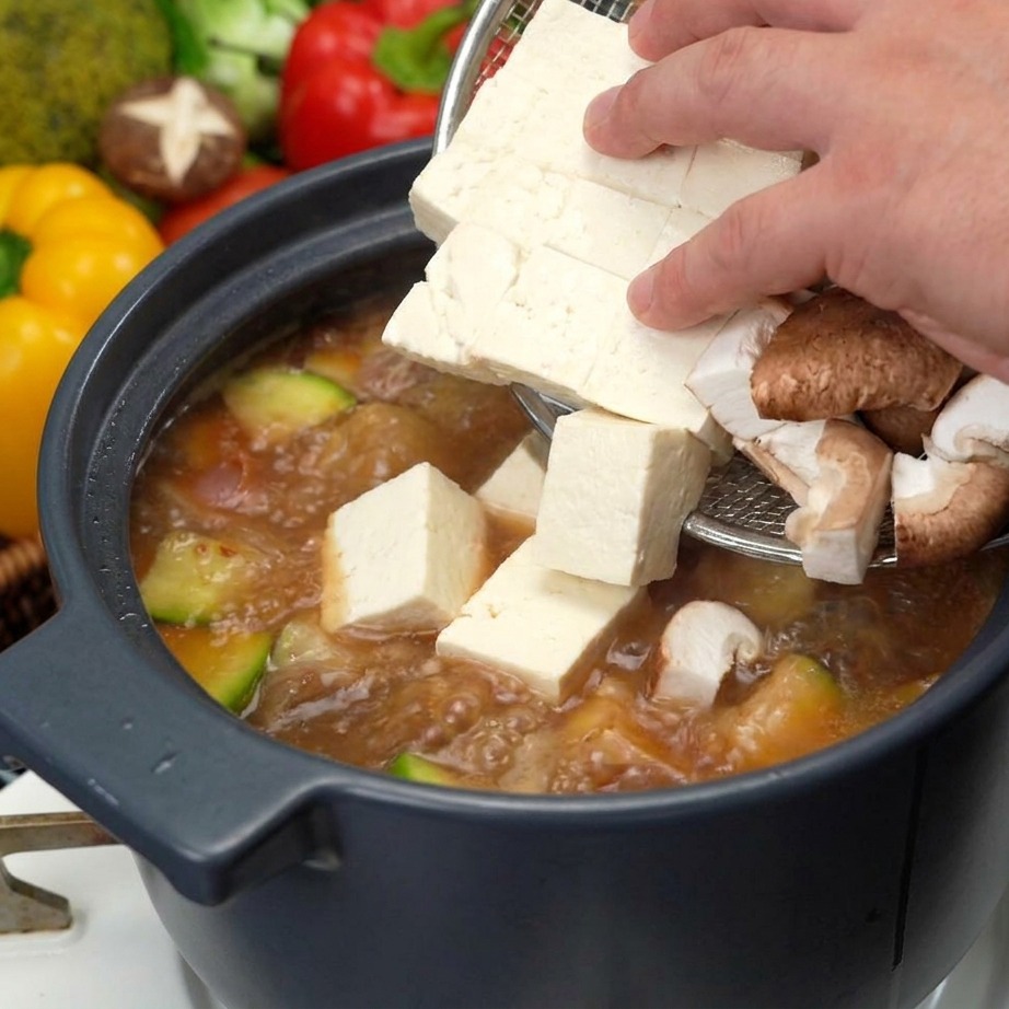 A hand using a sieve to add fresh white tofu cubes and sliced brown shiitake mushrooms into a pot of boiling soybean paste soup.