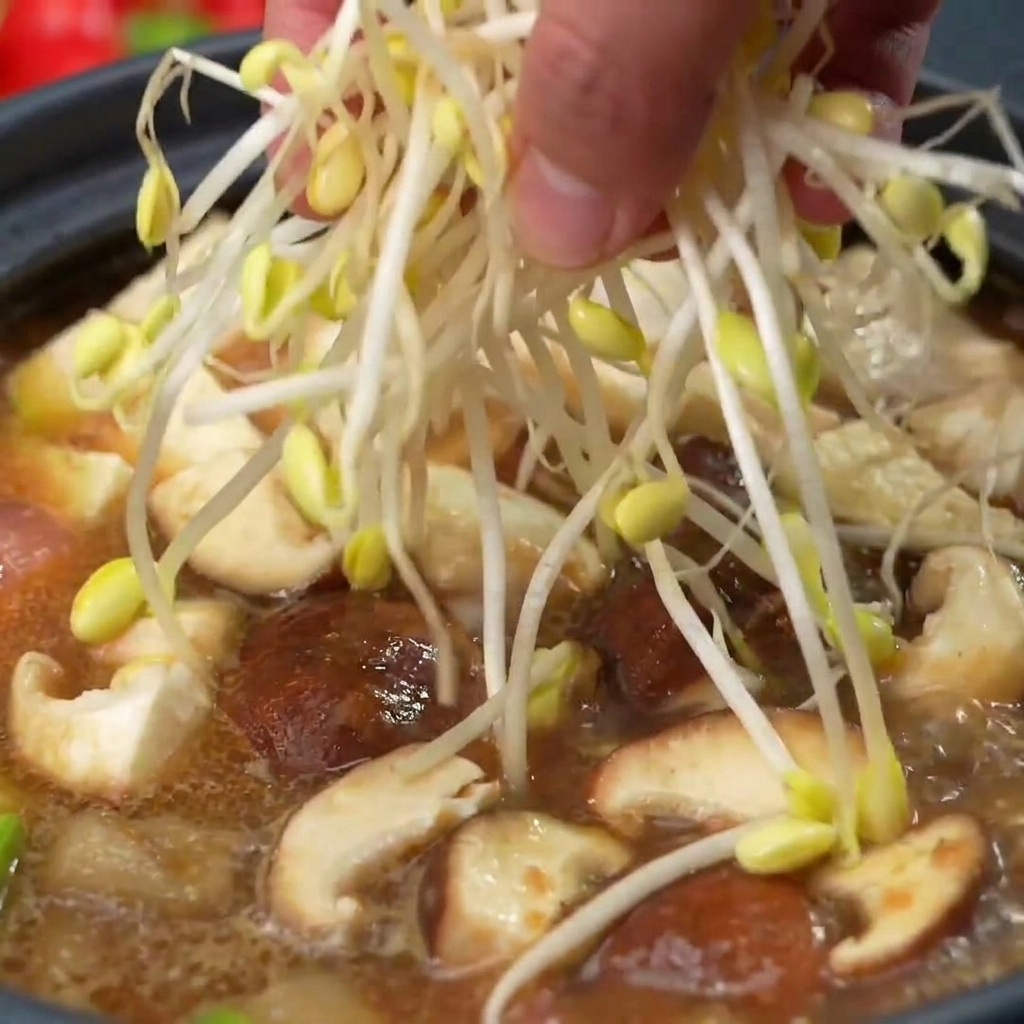 A hand placing a bundle of fresh, pale soy bean sprouts into the pot on top of mushrooms and other soup ingredients.