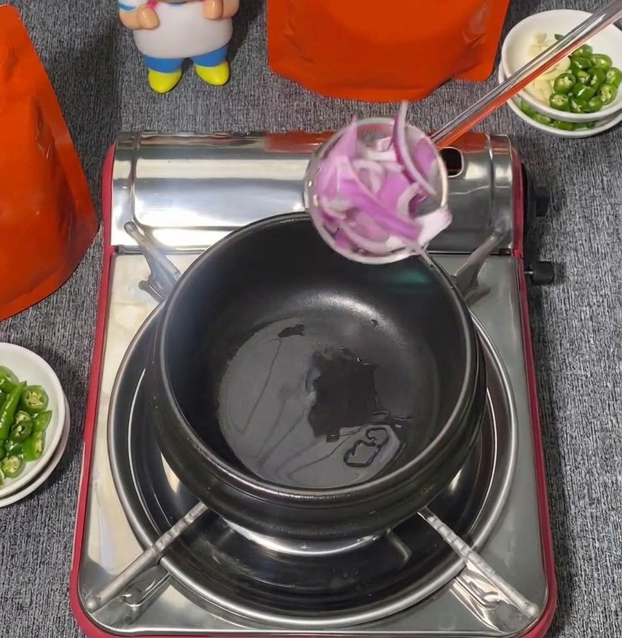 A ladle full of sliced red onions being lowered into a black ceramic cooking pot on a portable stove.
