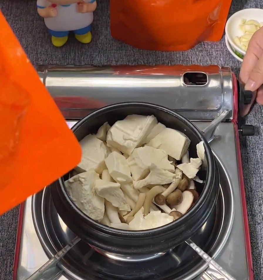 Large blocks of white soft tofu and shimeji mushrooms arranged in a black cooking pot.