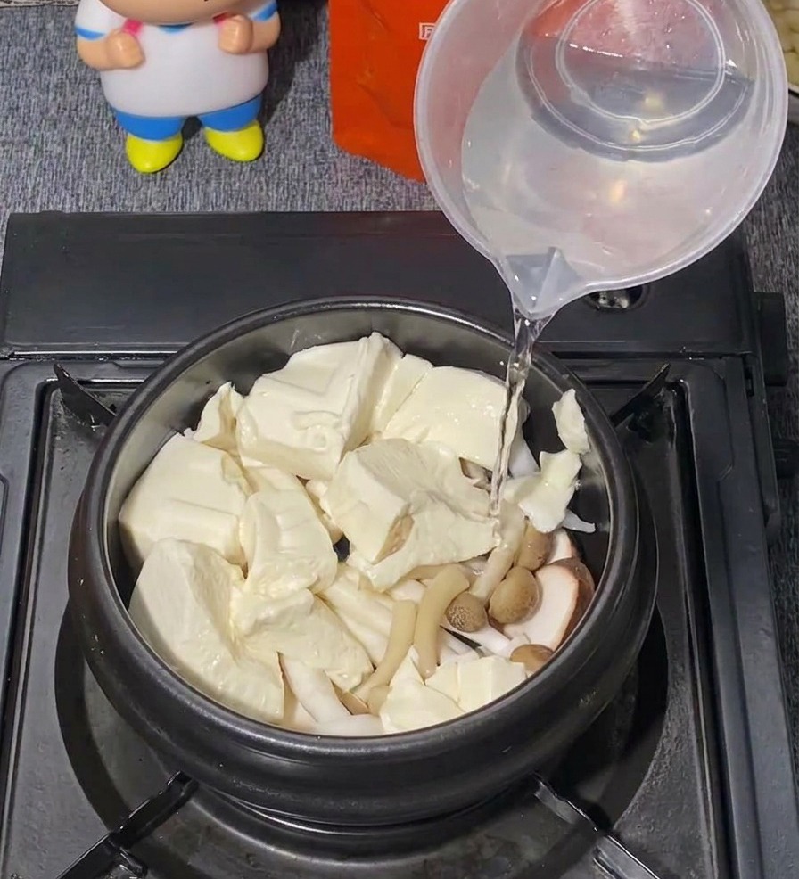 Clear water being poured from a plastic measuring cup into a black pot filled with tofu and mushrooms.
