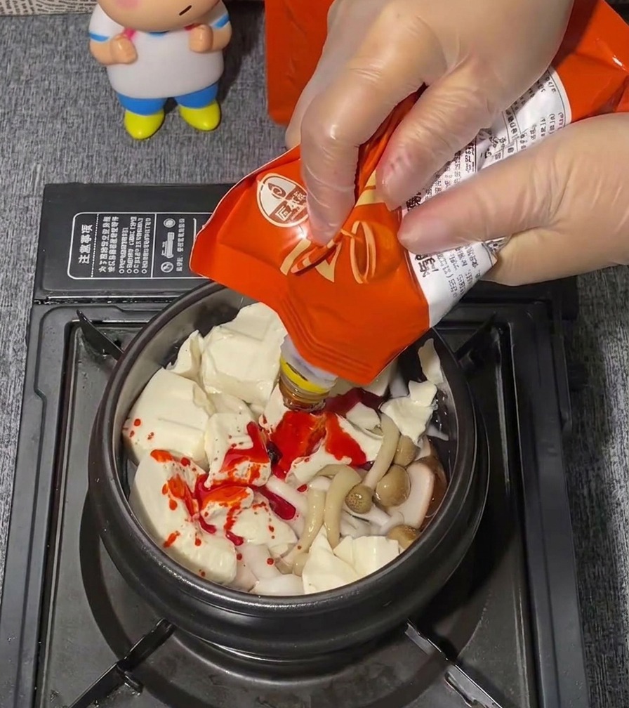 A hand in a clear plastic glove squeezing bright red seasoning paste from a pouch onto a bed of soft tofu.