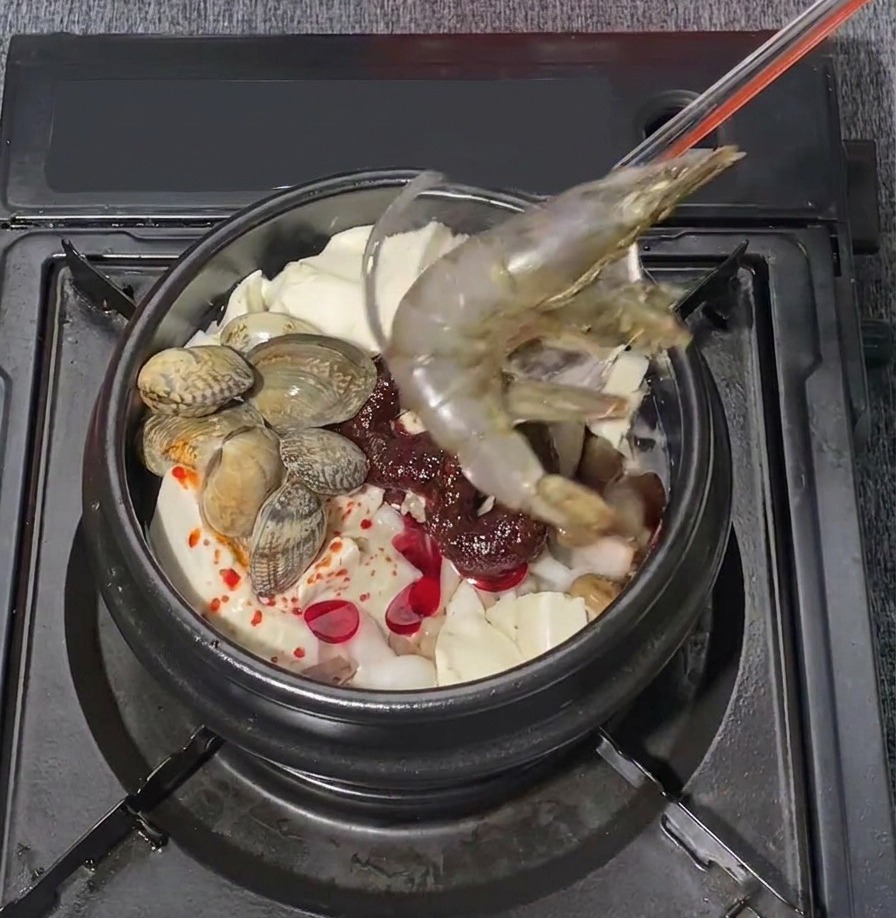 Two large raw prawns being carefully placed into a bubbling pot of Korean seafood soft tofu stew.