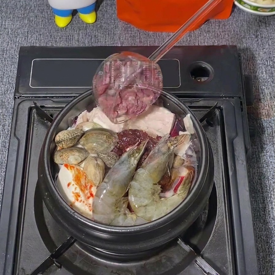 Thin slices of raw beef being added from a ladle onto a pot of Korean stew already containing prawns and clams.