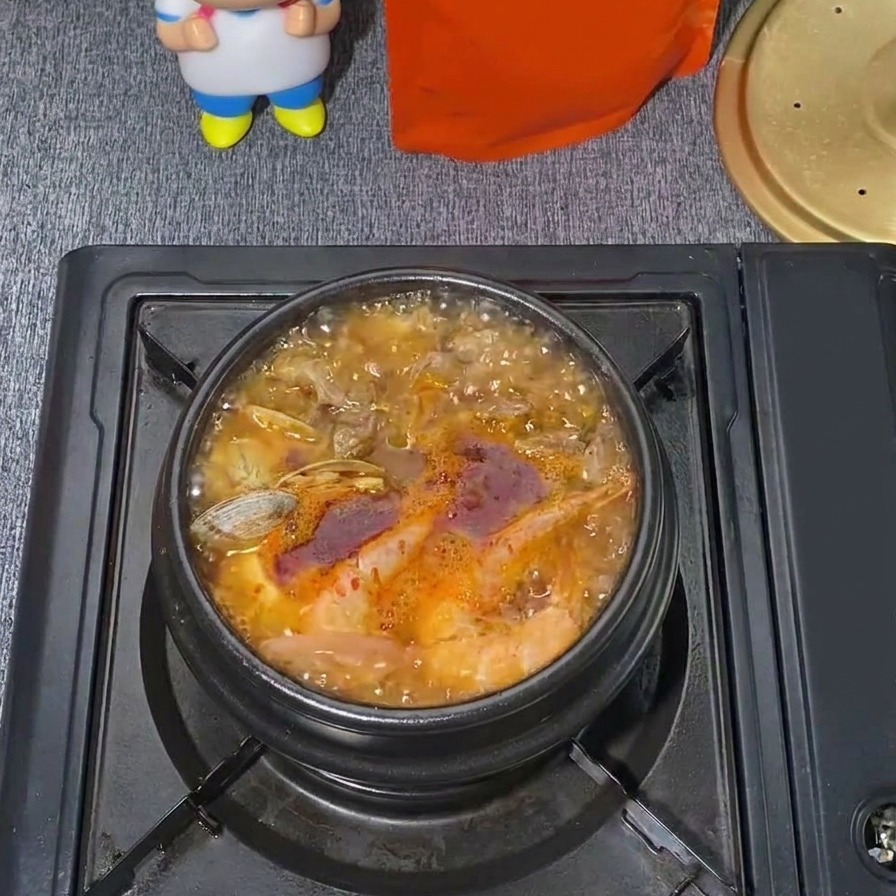 A bubbling pot of Korean seafood soft tofu stew with a vibrant red broth and visible clams and steam rising.