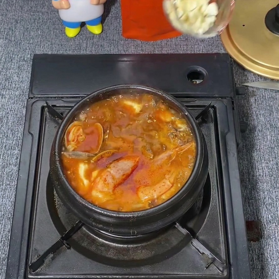 A clear ladle dropping a spoonful of minced white garlic into the center of a boiling, spicy red Korean stew.