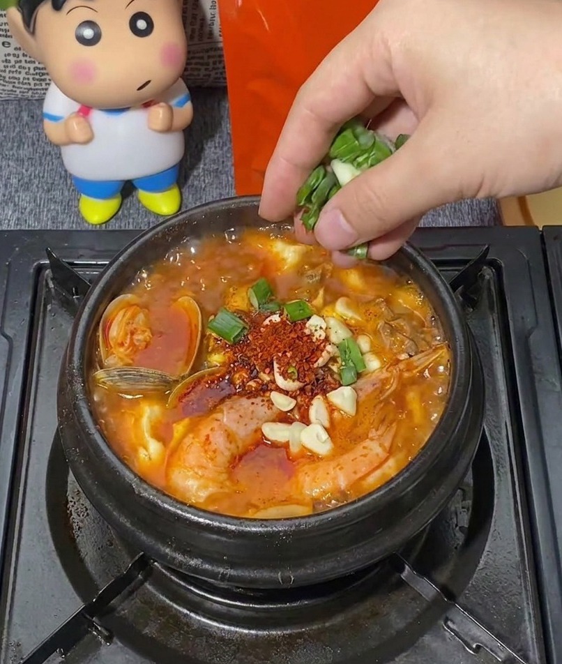 A hand sprinkling a bunch of chopped green scallions into a bubbling black stone pot of Korean seafood stew filled with shrimp, clams, and minced garlic.