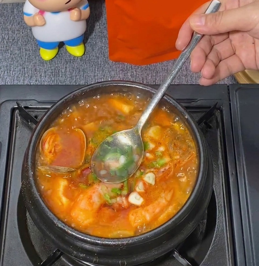 A metal spoon stirring chopped green scallions into a bubbling pot of red seafood soft tofu stew.