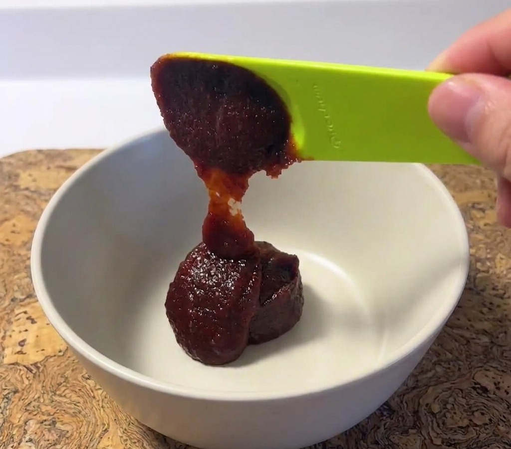 A dollop of thick dark red Korean chili paste being placed into a white ceramic mixing bowl with a green spoon