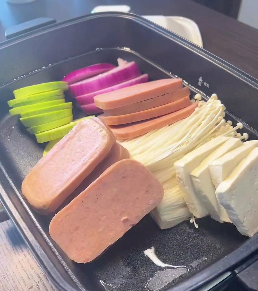 A black cooking pot filled with neatly arranged sections of red onion, green zucchini, luncheon meat, enoki mushrooms, and tofu blocks.