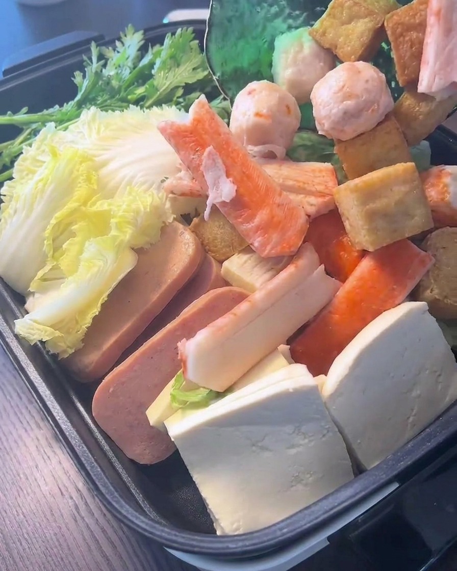 A variety of fish balls and crab sticks being added on top of fresh leafy greens and cabbage in the cooking pot.