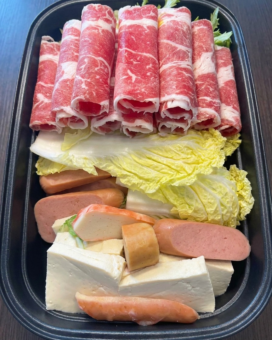 A hand placing several rolls of bright red, thinly sliced marbled beef onto the colorful arrangement of ingredients in the pot.