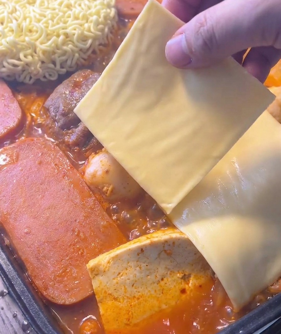 A hand placing a square slice of yellow cheese over instant noodles in a bubbling red stew