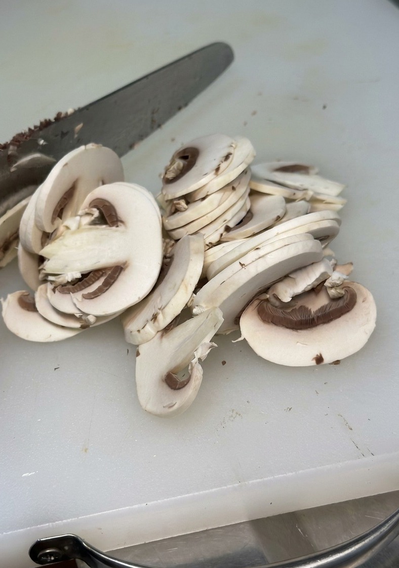 A close-up shot of a white mushroom being sliced into thin, even pieces with a metal knife on a white cutting board.