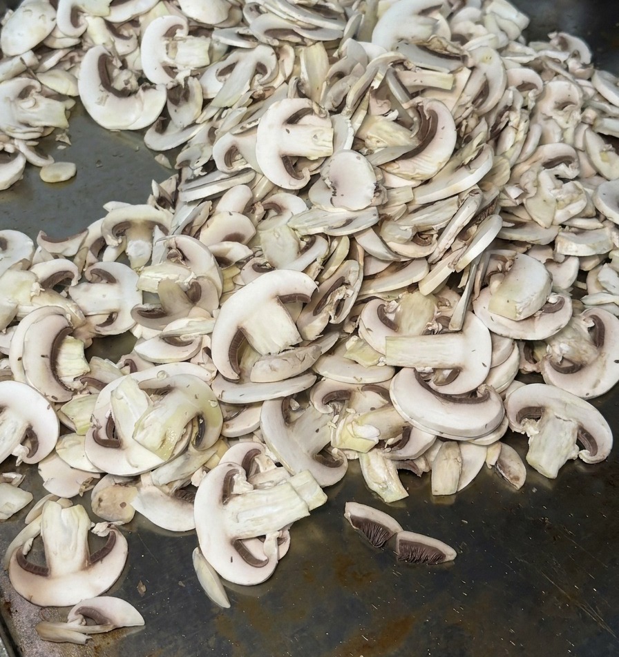 Thinly sliced mushrooms spread across a hot metal flat-top grill, releasing steam as they begin to cook.