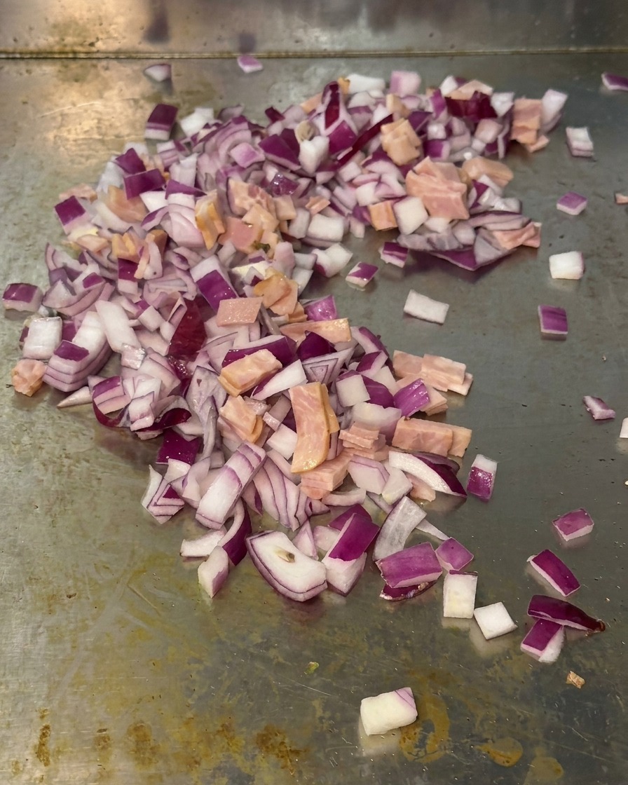 A mix of diced red onions and small pieces of bacon being sautéed together on a metal grill surface.
