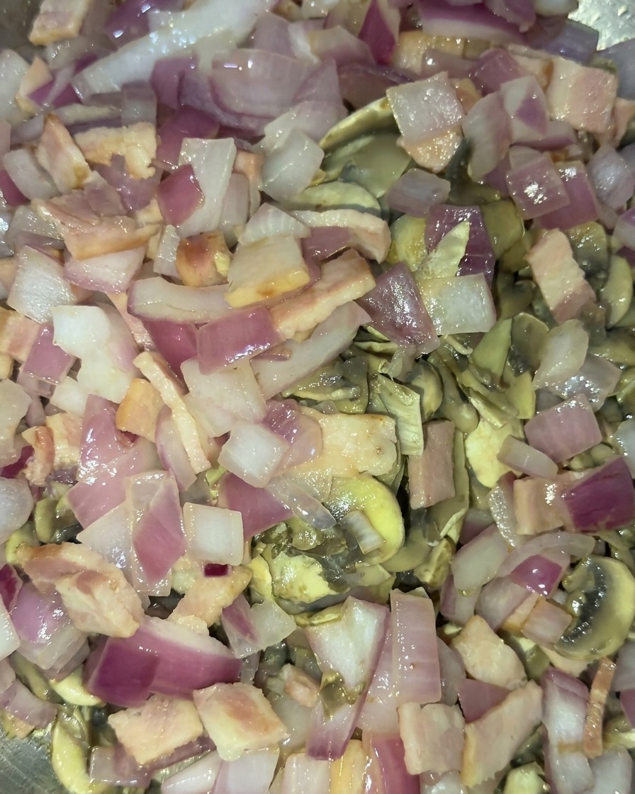 A combined mixture of golden sautéed mushrooms, translucent red onions, and cooked bacon bits being stir-fried.