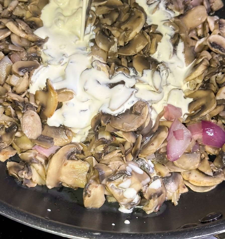 A stream of white heavy cream pouring from a blue carton into a pan filled with sautéed mushrooms and diced red onions.