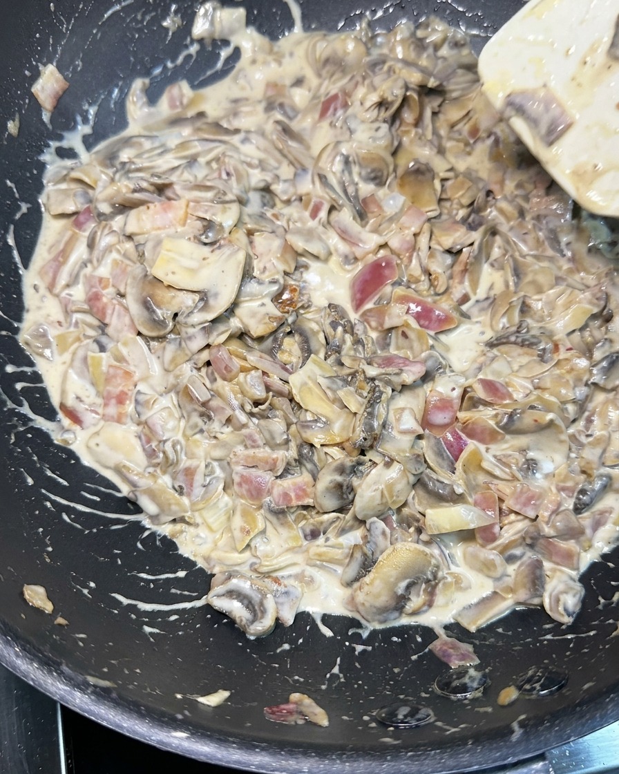 A white spatula stirring a thick, creamy sauce filled with sliced mushrooms, diced red onions, and bacon pieces in a black skillet.