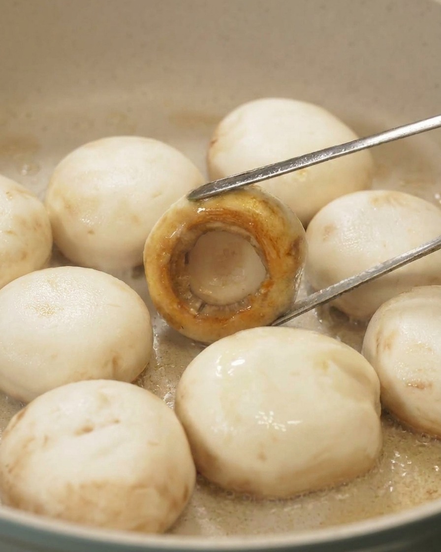 Tongs flipping a mushroom in a pan to reveal a perfectly caramelized, golden-brown surface on the cap.