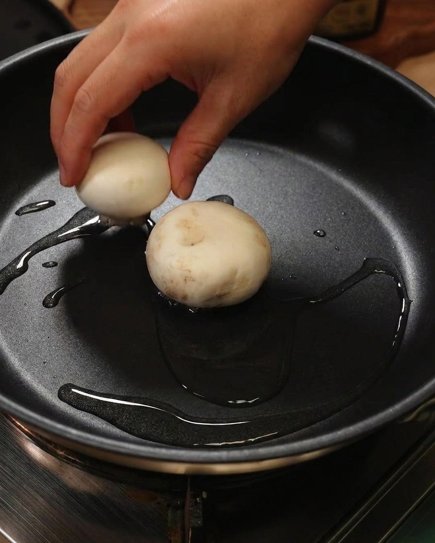 A hand placing a mushroom cap filled with shrimp paste face-down into a frying pan with oil.