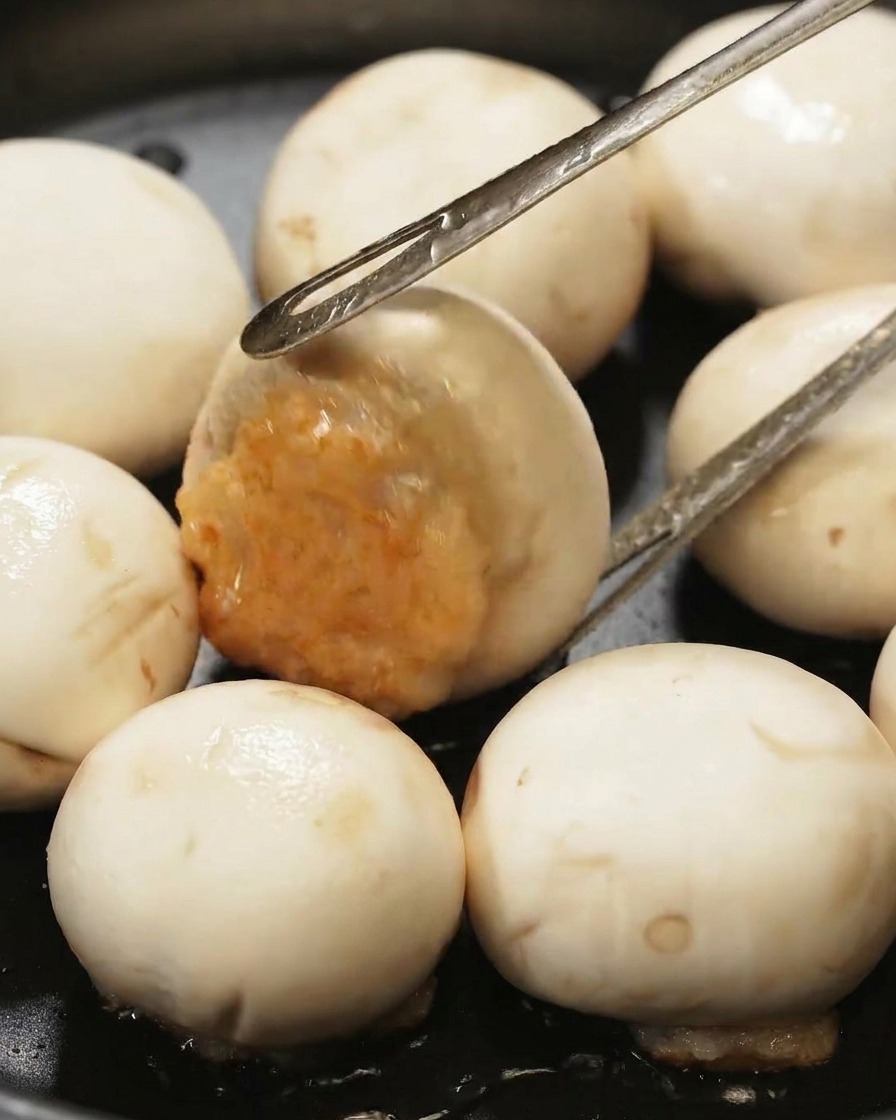 Metal tongs flipping a stuffed mushroom cap in a black pan to reveal the browned shrimp paste filling.