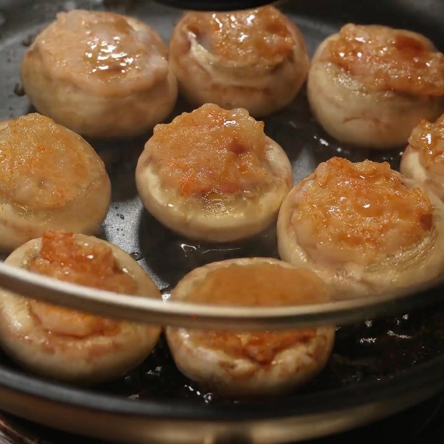 A person placing a clear glass lid onto a pan containing seasoned stuffed mushrooms.
