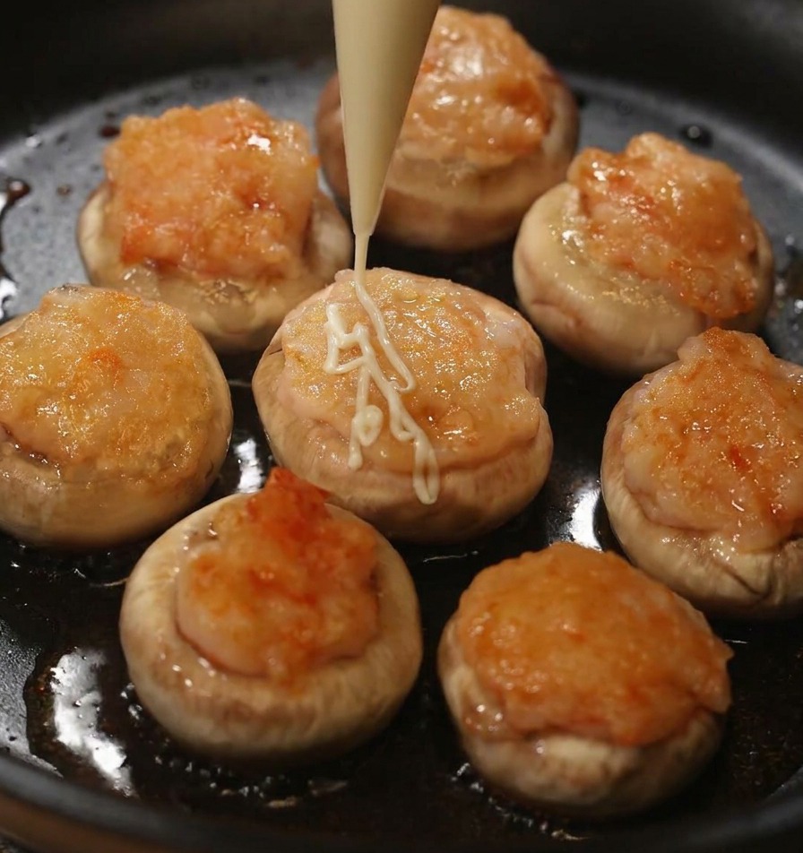 A person squeezing lines of white mayonnaise from a piping bag onto cooked mushrooms filled with shrimp paste.