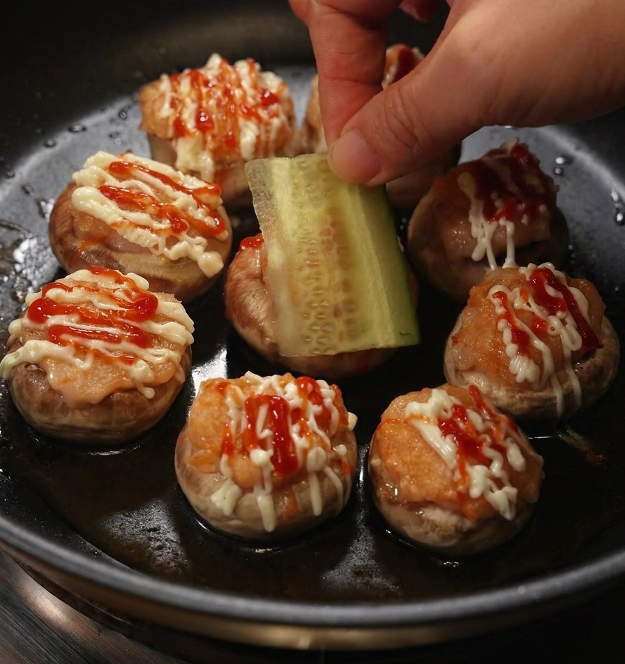 A hand placing a rectangular slice of fresh cucumber onto a stuffed mushroom burger topped with mayonnaise and ketchup.