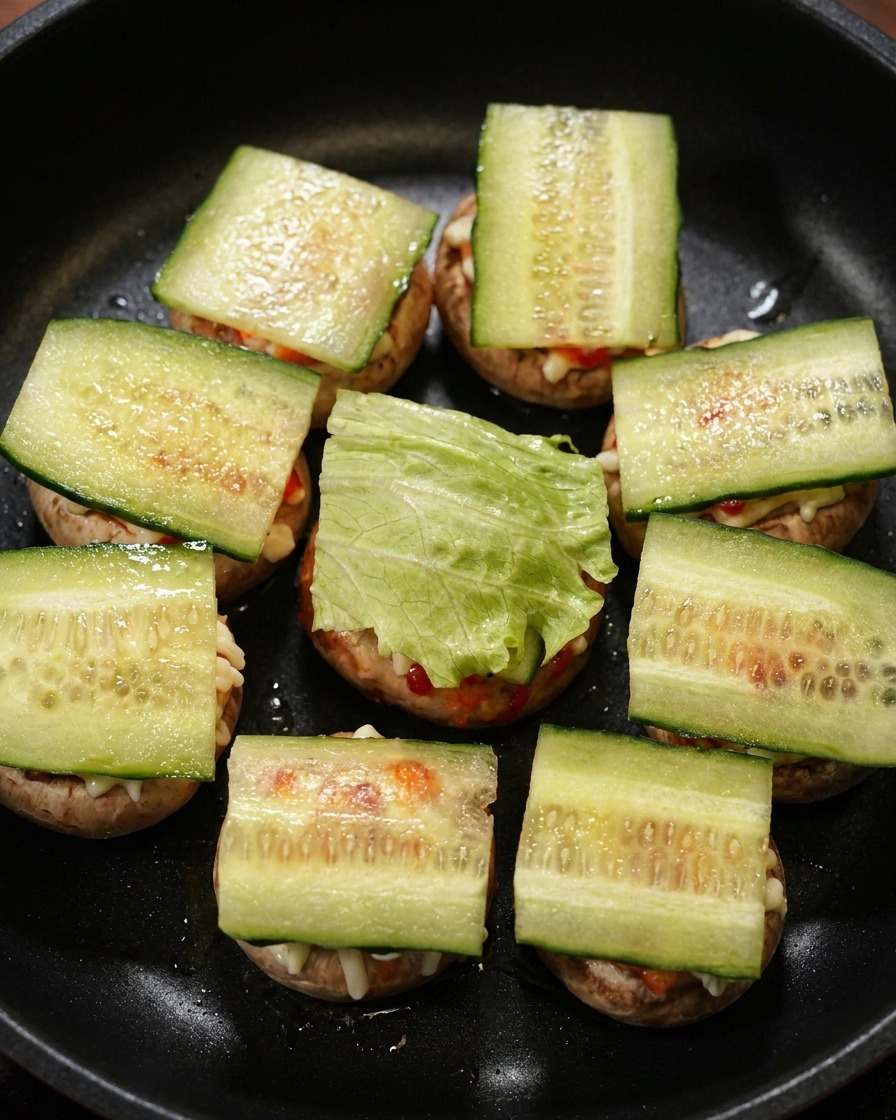 Slices of cucumber and green lettuce leaves layered on top of mini mushroom burgers in a black skillet.