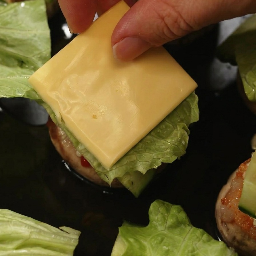 A hand placing a square slice of yellow cheese on top of lettuce and cucumber inside a mushroom burger.