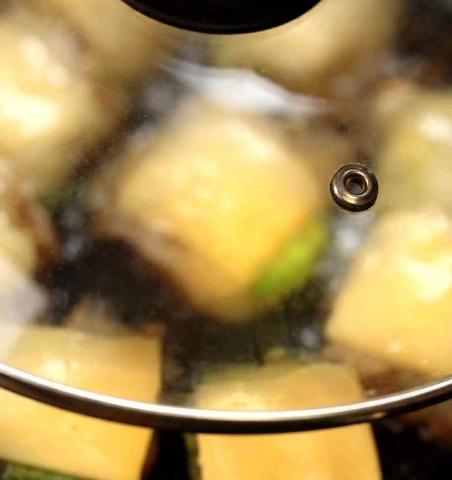 A glass lid covering a pan where cheese slices are starting to melt over mushroom burgers.