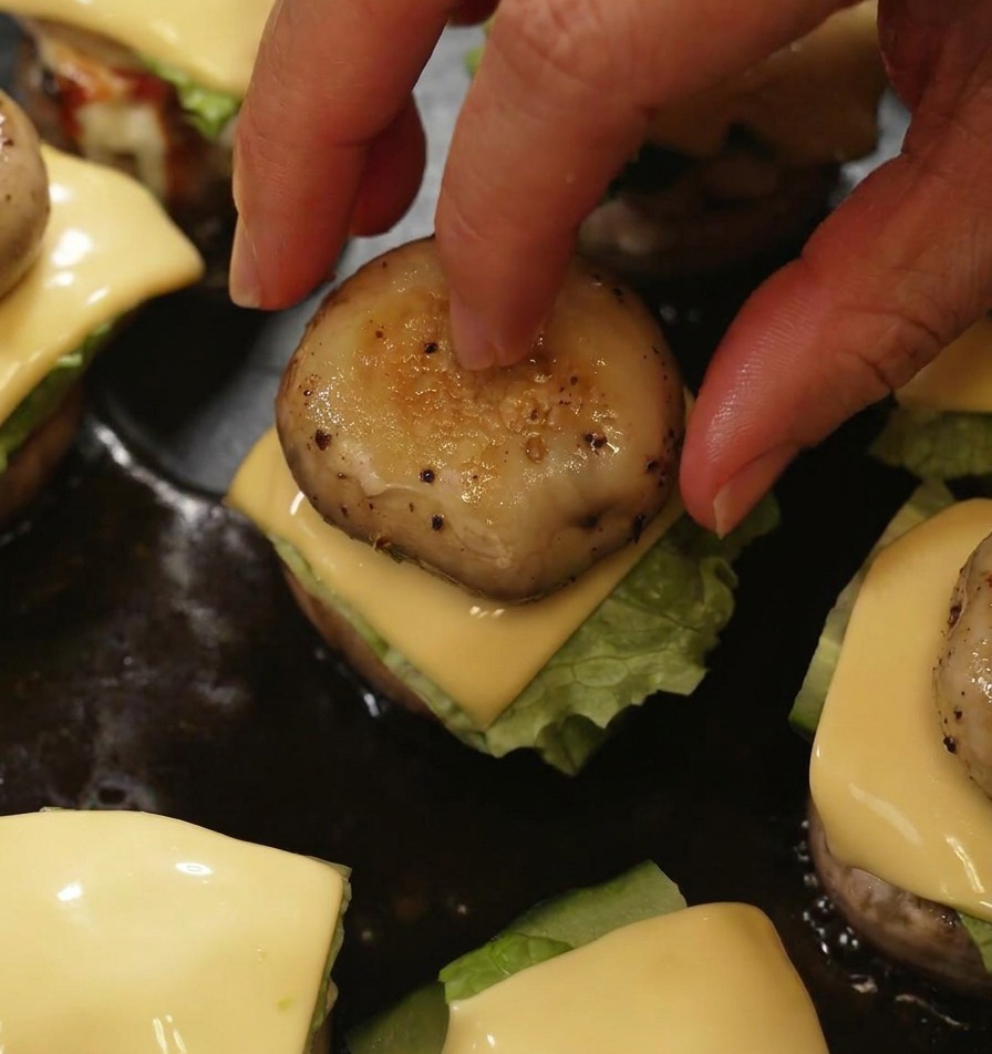 A hand placing a grilled mushroom cap onto a mini burger to complete the assembly in the pan.