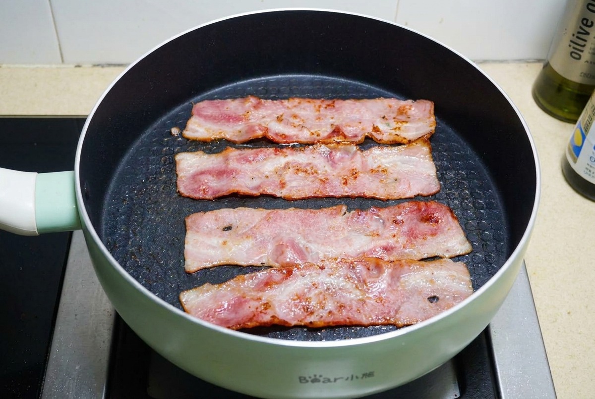 Four strips of bacon frying in a black non-stick pan until the fat is bubbling and the meat is golden brown and crispy.