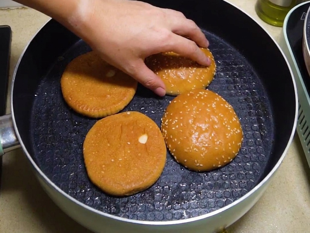 Four burger bun halves being toasted face-down in a pan with a hand reaching in to adjust their position.